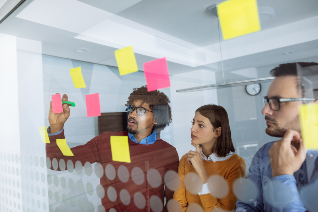 Three colleagues collaborating during a brainstorming session, placing colorful sticky notes on a glass partition wall